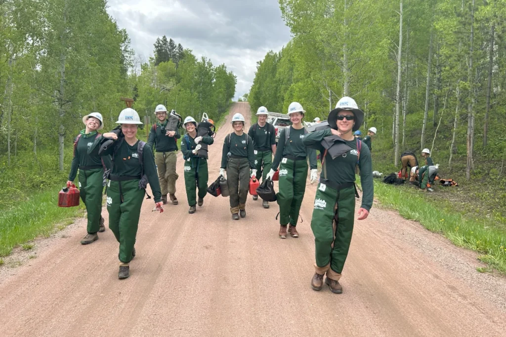 A group of people in safety helmets and uniforms walk down a dirt road carrying equipment, with trees and a cloudy sky in the background.
