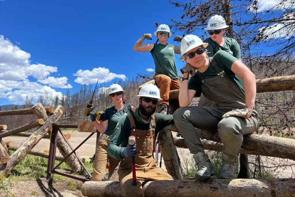 Five workers in hard hats and work clothes pose on a wooden fence in a rural outdoor area, with tools and trees in the background under a partly cloudy sky.