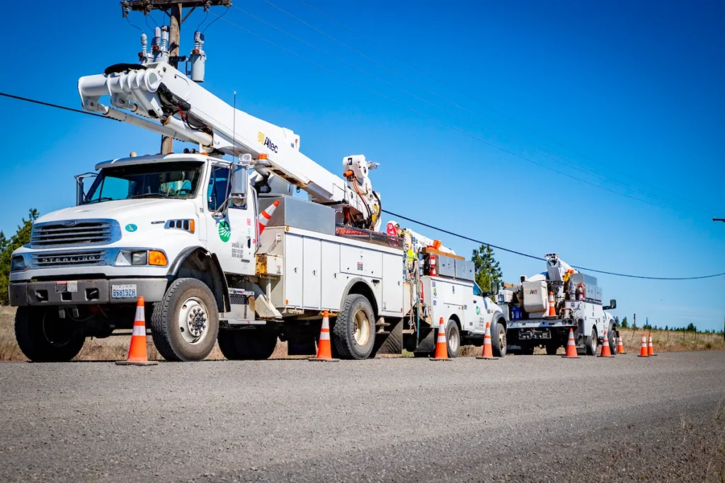 Three utility trucks with raised booms are parked on a roadside, surrounded by orange safety cones, under a clear blue sky.