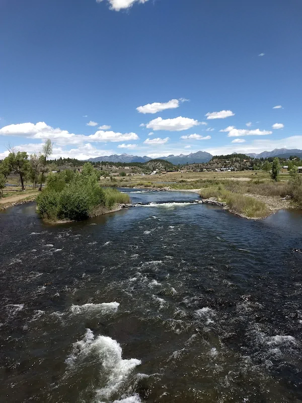 A river flows under a partly cloudy sky with grass, shrubs, and distant mountains visible along the horizon.
