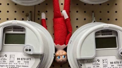 An elf doll hanging upside down between electric meters on a pegboard wall.