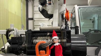 A small elf doll in red sits on a large hook attached to machinery inside an industrial room.