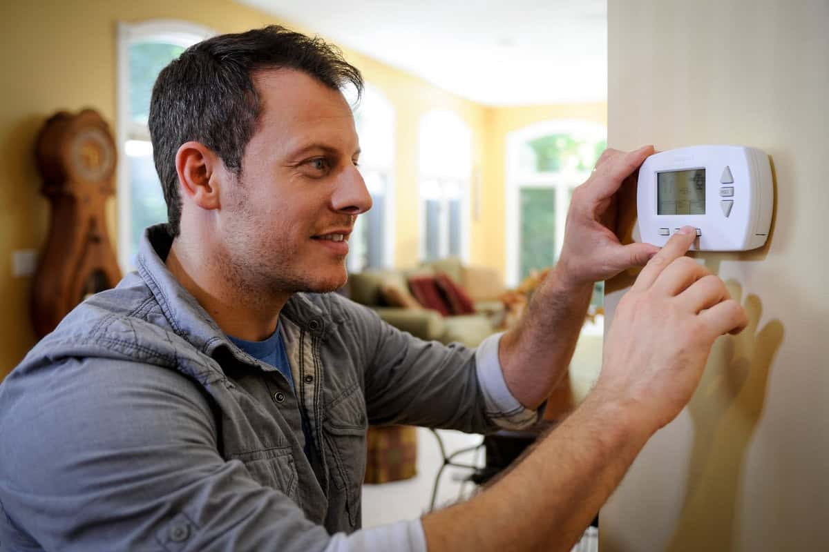 A man in a blue shirt adjusts a thermostat on a wall in a brightly lit living room with a grandfather clock in the background.