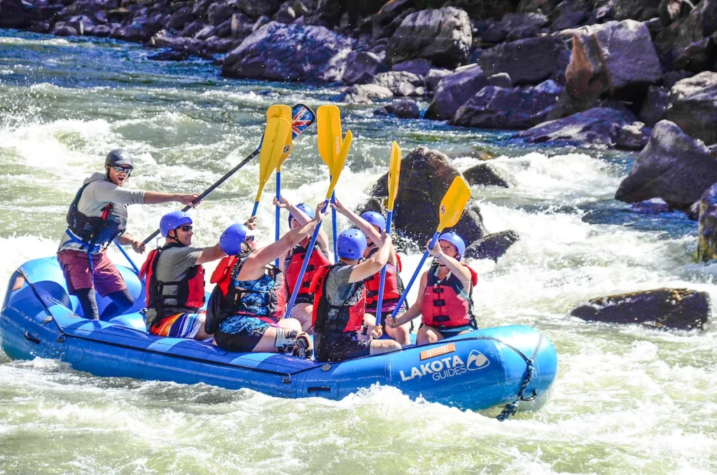 A group of people wearing life jackets and helmets paddle a blue inflatable raft down a rocky, fast-moving river.