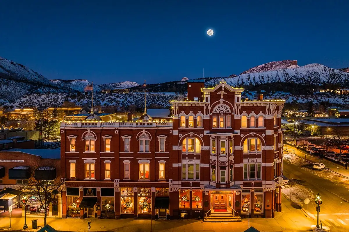 Historic red-brick building illuminated at night, with snow-covered mountains and a full moon in the background.
