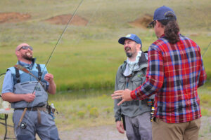 Three men outdoors laughing while holding fishing rods near a grassy area.