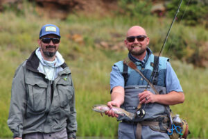 Two men outdoors, one holding a fishing rod and a small fish. Both are wearing fishing gear and sunglasses, standing on grassy terrain.