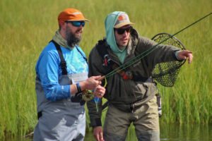 Two men in outdoor clothing fish in a grassy area. One man holds a fishing rod, while the other points, wearing a net on his back.