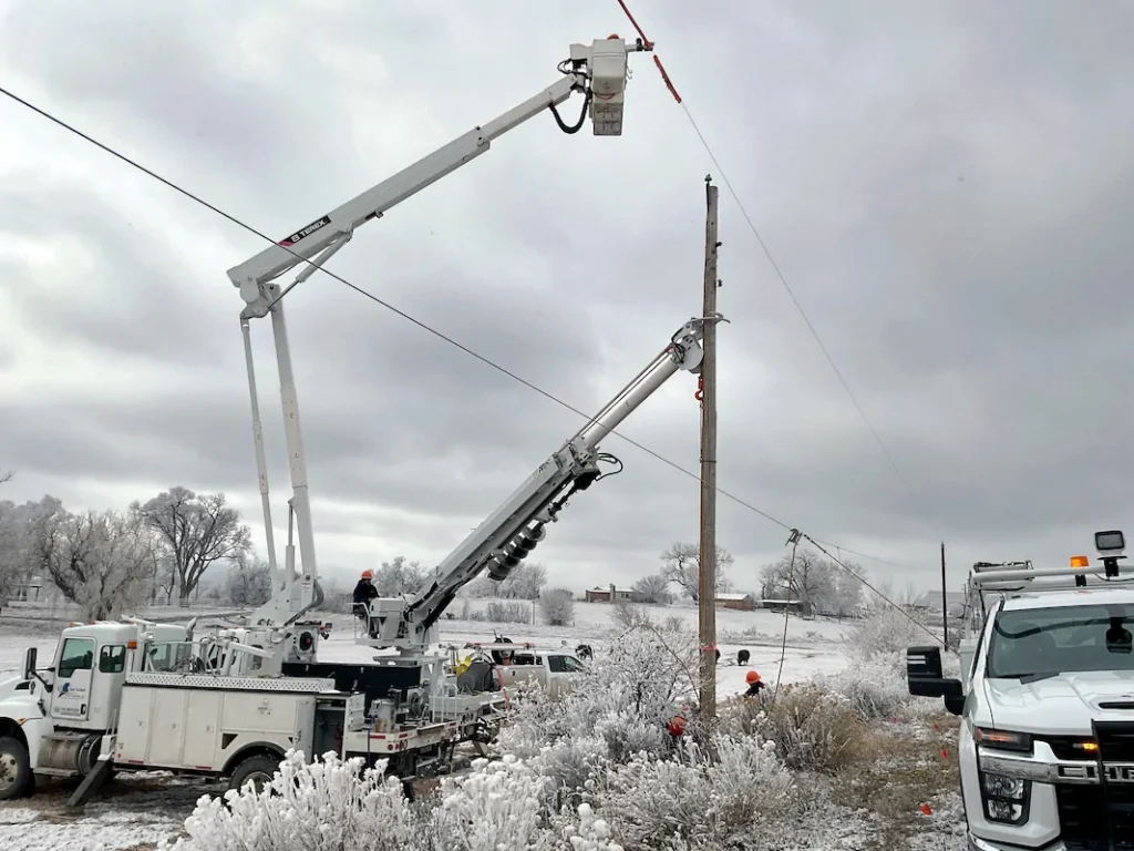Utility workers in bucket trucks repair power lines on a snowy, overcast day, surrounded by frosted trees and several service vehicles.