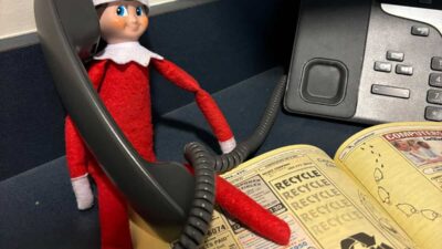 A toy elf in a red outfit is holding a phone receiver while sitting on a table next to an open phone book and a telephone.