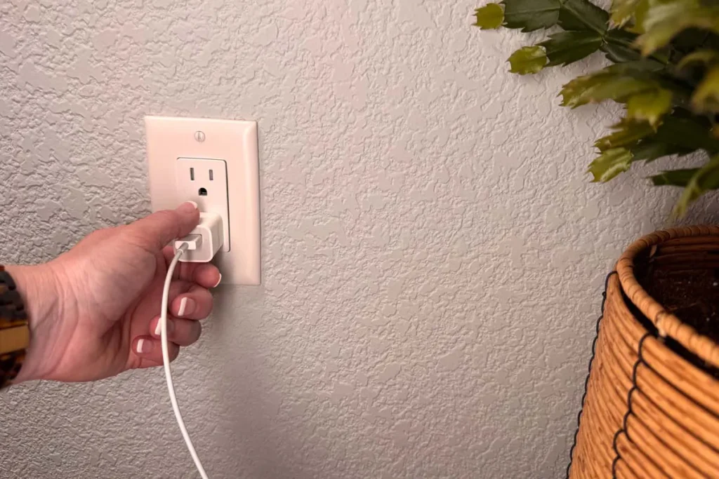 A hand is plugging a white power adapter into a wall electrical outlet near a wicker basket planter with a green plant.