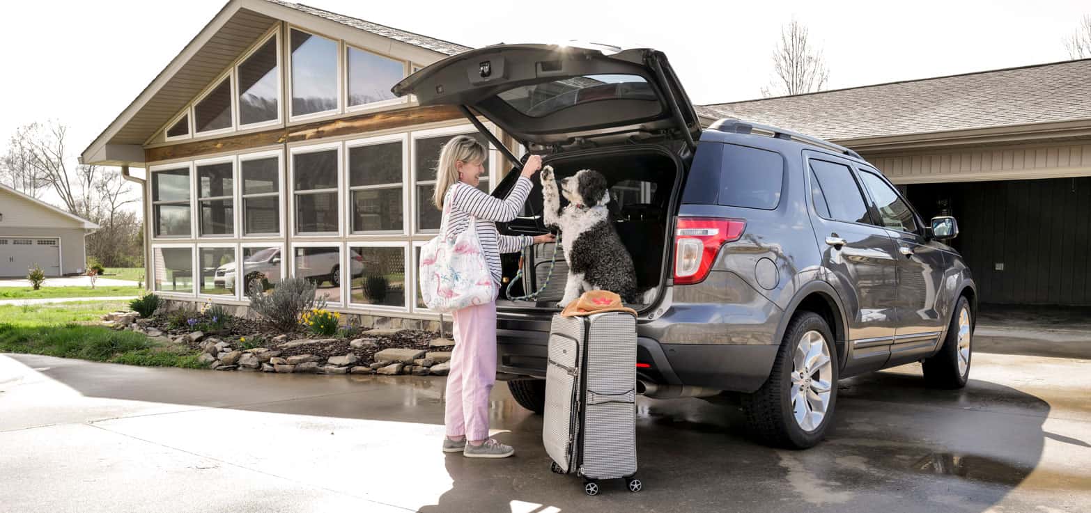 A person places luggage in the back of an SUV while a dog sits inside. A house with large windows is in the background.