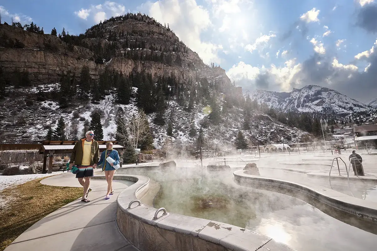 Two people in swimsuits and towels walk beside a steaming outdoor hot spring pool, with snow-covered mountains and pine trees in the background.
