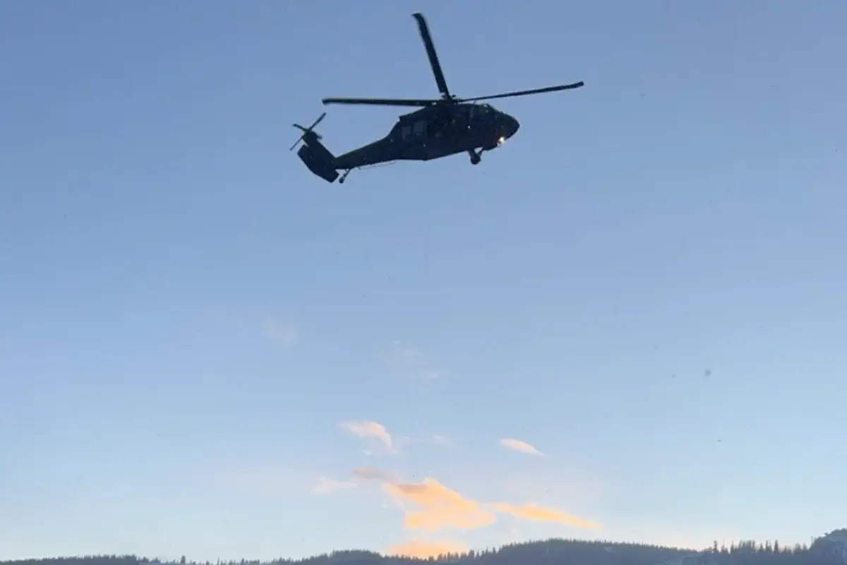 The Next Generation of Search and Rescue A helicopter flies overhead against a clear blue sky, with distant hills and a small orange cloud visible near the horizon.
