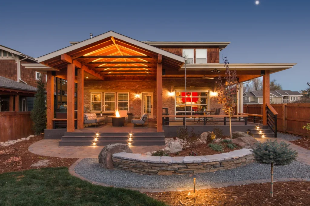 A modern backyard patio with covered seating area, fire pit, outdoor furniture, stone accents, and garden landscaping, illuminated by evening lighting.