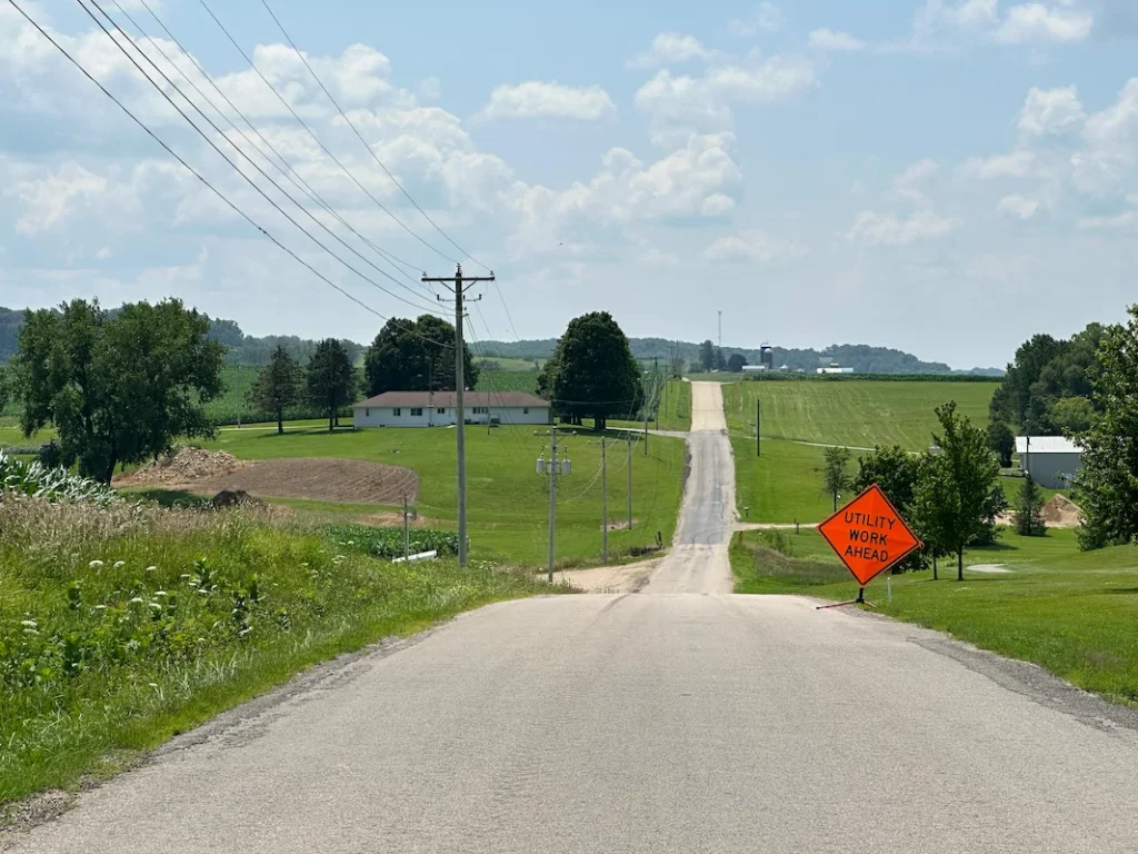 A rural road stretches into the distance with a "Utility Work Ahead" sign on the right; fields, houses, and power lines are visible under a partly cloudy sky.